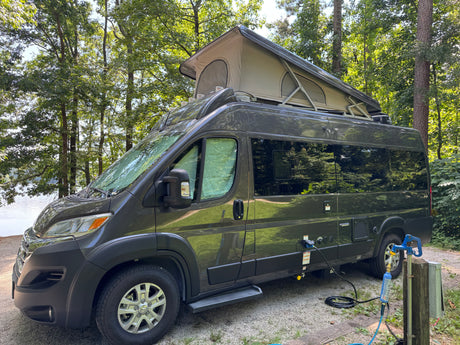 Modern gray rv rental camper van with a pop-top roof, parked at a scenic lakeside campsite and hooked up to utilities.