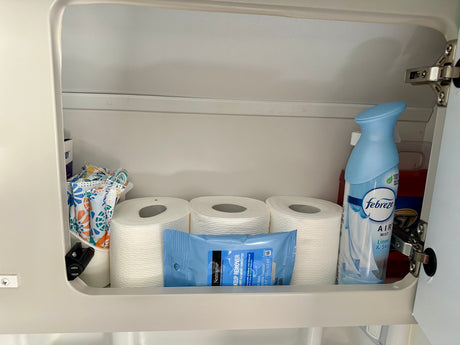A neatly organized RV rental cabinet holds toilet paper, Neutrogena wipes, and Febreze air freshener for personal care needs.