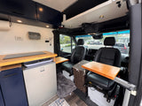 This customized RV rental interior features a kitchen counter, refrigerator, swiveling front seats with pull-out tables, and overhead storage.