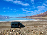A dark gray RV rental van parked beside a vast, reflective salt flat in a desert valley, with distant mountains under a blue sky.