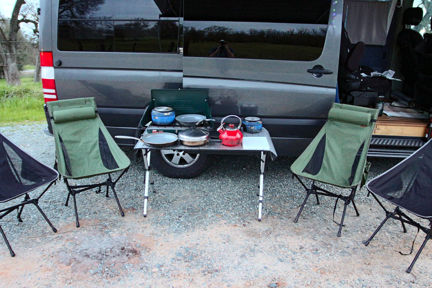 Outdoor cooking scene with a gray RV rental van, portable stove, pans, kettle, and folding chairs on a gravel area.