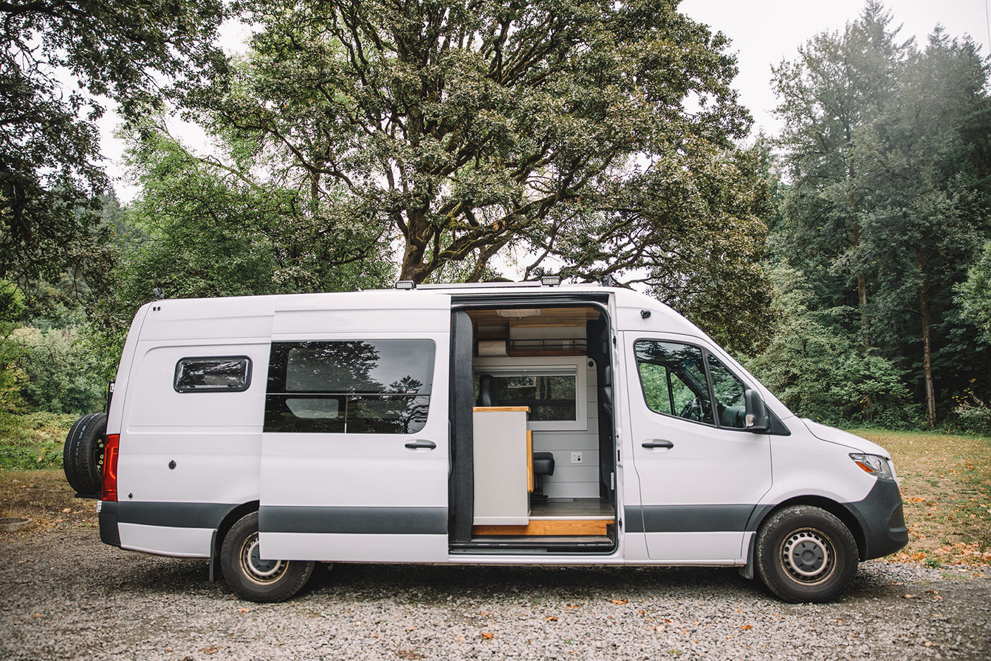 A white camper van rv rental with its side door open, revealing a built-out interior, parked in a treed natural setting.