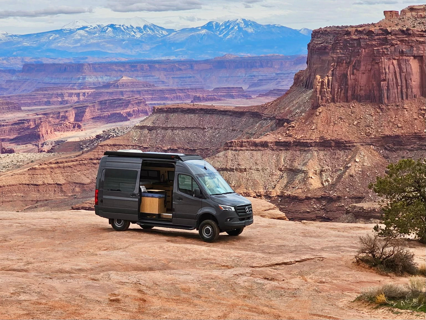A gray rv rental camper van overlooks a stunning, layered canyon landscape with snow-capped mountains in the distance.