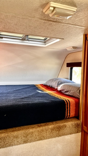  and a wall heater under a window.|Inside an rv rental: inviting dining area & kitchen with a stunning coastal view outside the large window. Ready for adventure!|Relaxed woman reading in an RV rental dinette overlooking a scenic beach. Perfect for your next travel adventure.|RV rental interior: Screen door shows a beach view. Features include a kitchen sink — photo 9