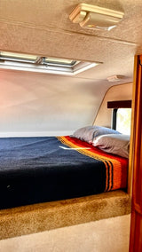 Cozy RV rental sleeping area features a bed with pillows, a striped blanket, and an overhead skylight for travel comfort.