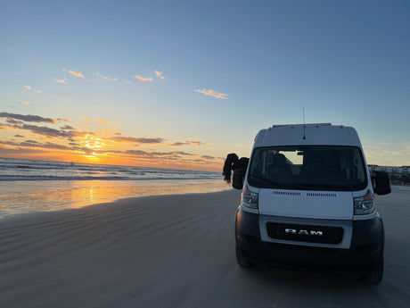 White rv rental van on a beach at sunset. Person leans out to see golden ocean & sky views.