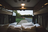 Inside a cozy RV rental: comfy bed, open doors revealing a scenic road, trees, and shed. Ready for adventure!