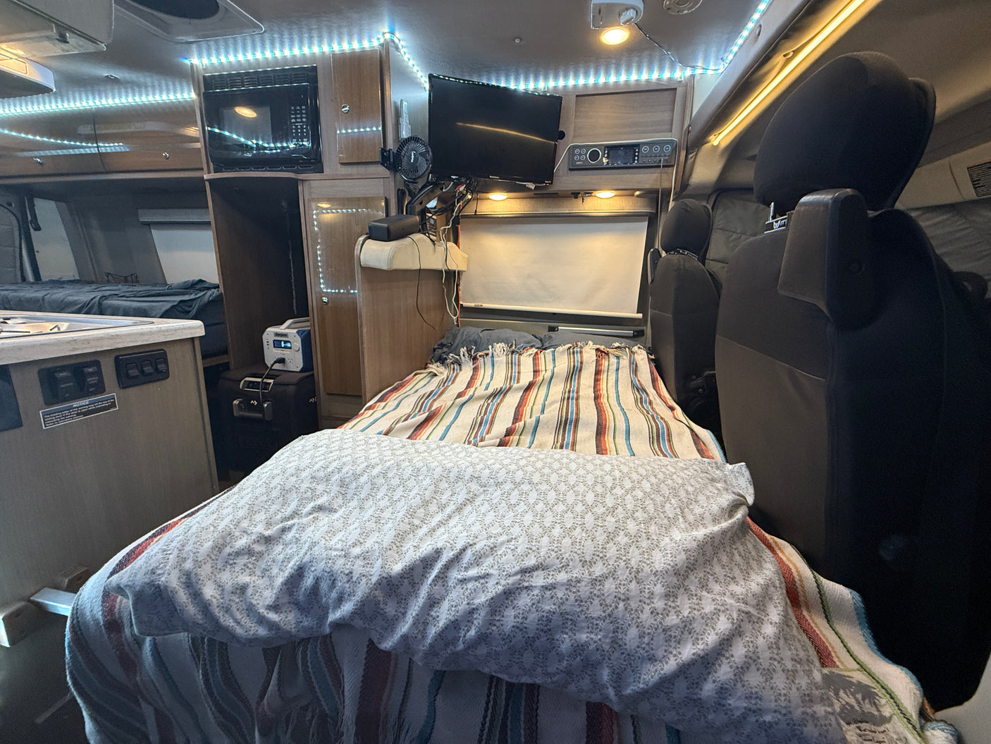 Interior of an RV rental showing a bed with striped and patterned covers, a compact kitchen, overhead microwave, TV, and front seats.
