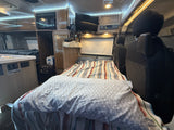 Interior of an RV rental showing a bed with striped and patterned covers, a compact kitchen, overhead microwave, TV, and front seats.