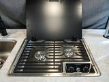 RV rental kitchen showcasing a sleek Dometic 2-burner stovetop with a black lid raised, next to a stainless steel sink.