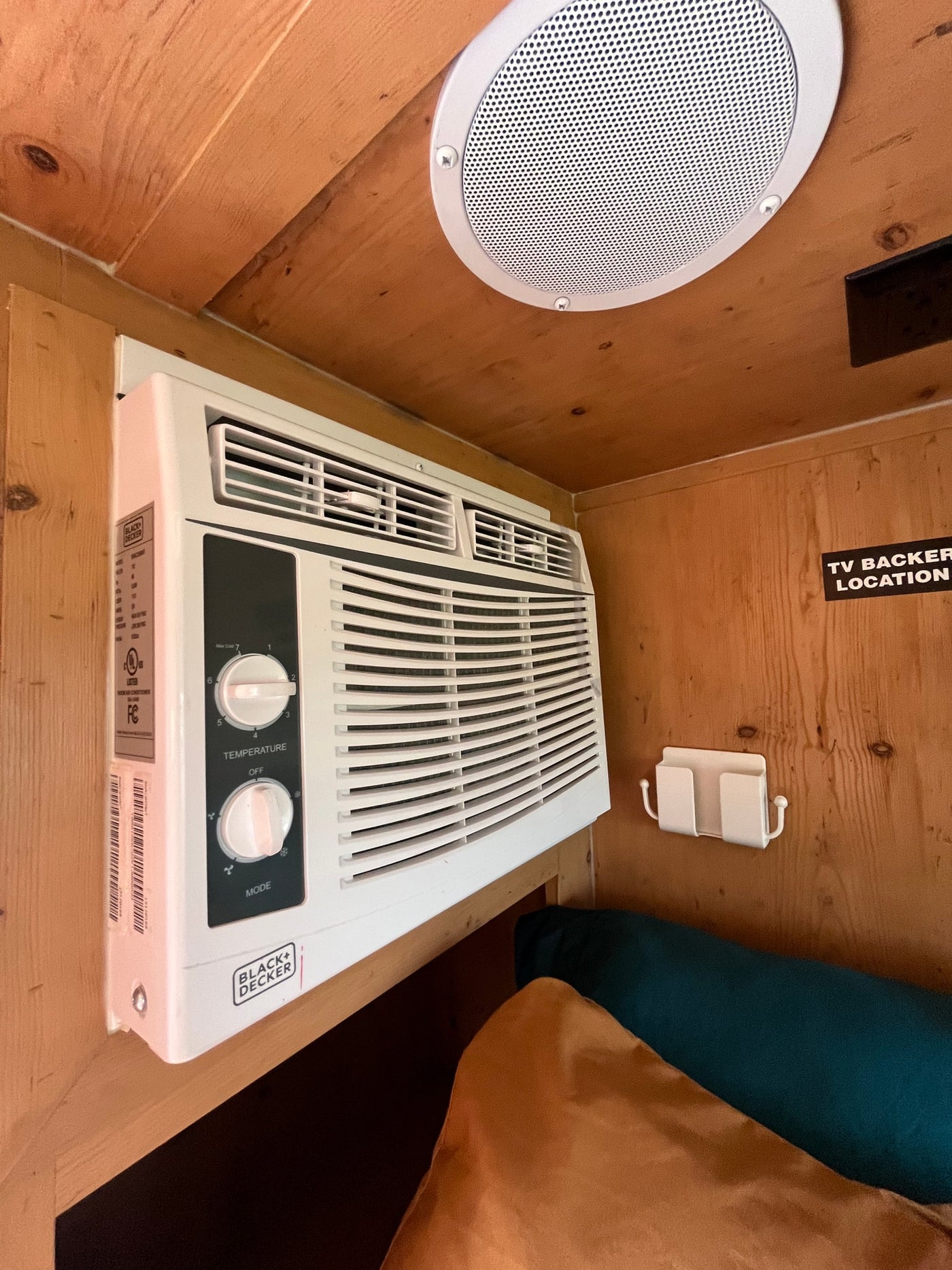 RV rental cabin: Wall-mounted Black+Decker AC unit, ceiling speaker, and wooden interior details, including a "TV BACKER LOCATION" sign. — photo 9
