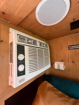 RV rental cabin: Wall-mounted Black+Decker AC unit, ceiling speaker, and wooden interior details, including a "TV BACKER LOCATION" sign.
