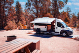RV rental campervan parked at a scenic campsite with an extended awning, chairs, picnic table, and fire pit setup.