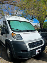 White Ram ProMaster van with a green sunshade, parked under leafy trees. Ideal for an RV rental adventure.