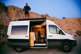 Adventurer on an rv rental camper van roof in a desert landscape. Its side door is open, revealing a cozy mobile home interior.