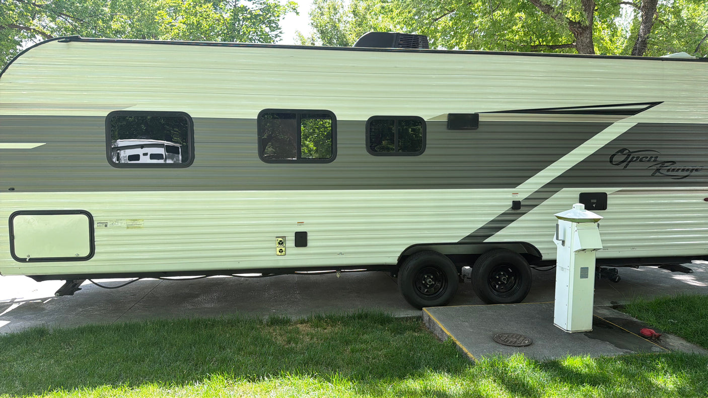 This rv rental, an Open Range travel trailer, is parked at a tree-lined campsite with utility hookups for power and water.