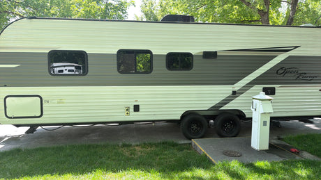 This rv rental, an Open Range travel trailer, is parked at a tree-lined campsite with utility hookups for power and water.