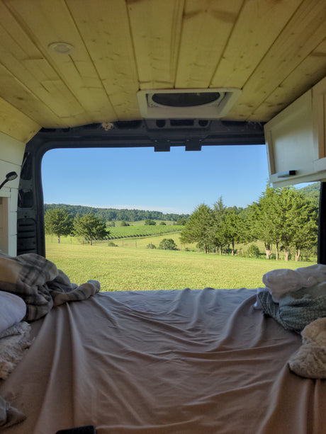 Experience an rv rental! Cozy camper van bed with a stunning view of a green vineyard, hills, and clear sky. Relaxing travel.