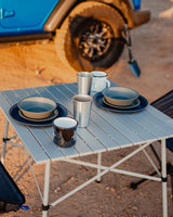 Portable table with dinnerware set for two, beside a blue vehicle in a desert landscape. Ideal for your RV rental trip.