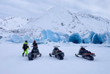 Three snowmobiles and two adventurers face a vibrant blue glacier and snowy mountains. Discover winter with an RV rental!
