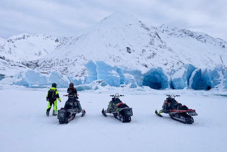 Three snowmobiles and two adventurers face a vibrant blue glacier and snowy mountains. Discover winter with an RV rental!