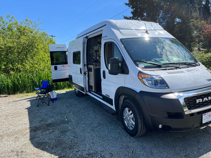 White Solis camper van RV rental parked on gravel with open door and chairs, set up for a sunny outdoor day.