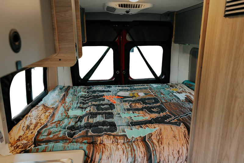 RV rental interior: a comfortable bed with a colorful patterned comforter, visible bright windows at the back. — photo 6
