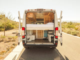 Rear view of an RV rental with open doors, revealing a wooden interior with elevated bed and utility space for van life.