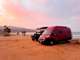 Red rv rental van & black SUV with rooftop tent parked at a sandy beach during a vibrant pink sunset.