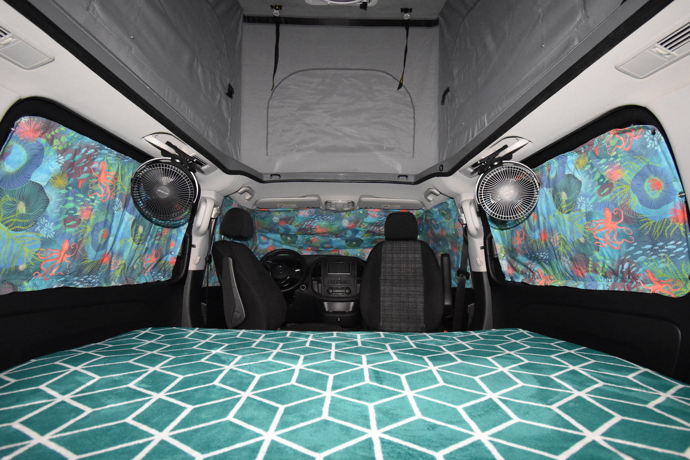 Inside an RV rental: a cozy camper van with a geometric-patterned bed, pop-top roof, and colorful window curtains.