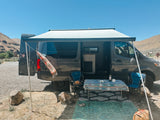 This rv rental camper van boasts an extended awning, table, chairs, and rugs, creating a comfy outdoor setup in a sunny desert landscape.