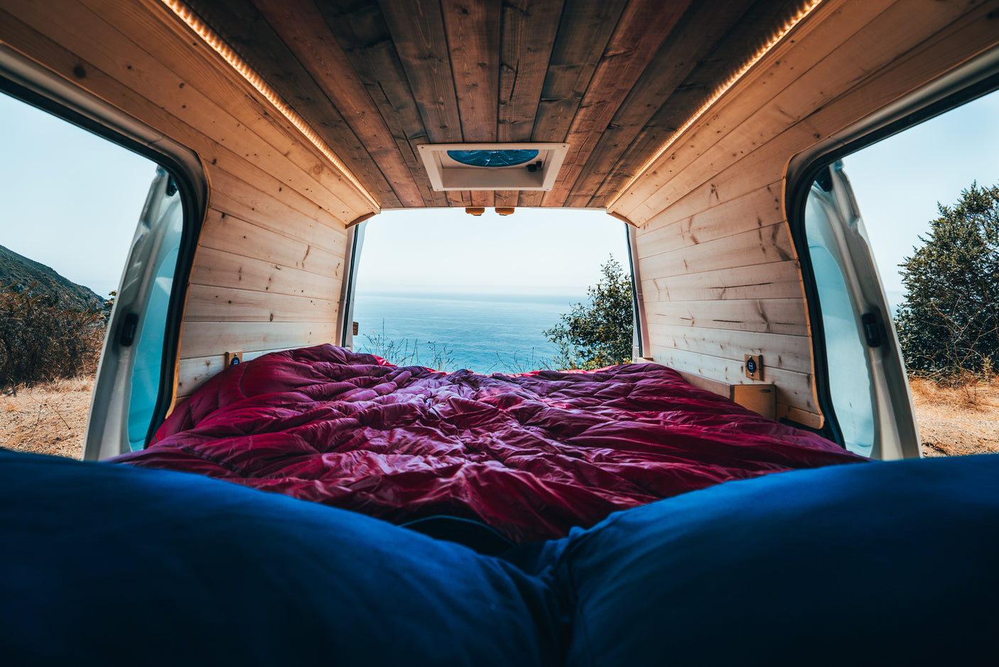 Scenic rv rental: Inside a wood-paneled van, a red bed and person's legs face an expansive ocean view, framed by open doors.