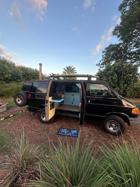 This black camper van, an RV rental, with surfboards on top, doors open revealing its custom interior. Parked for adventure.