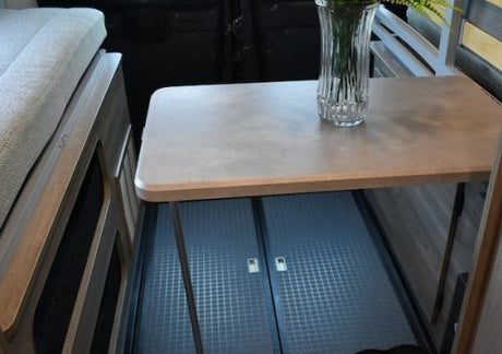 This RV rental's compact interior features a dining table with a vase, offering under-table storage, next to a cushioned bed.