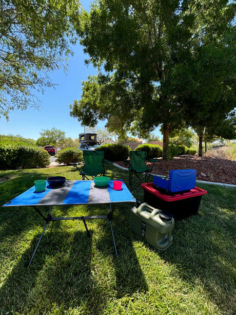 Sunny park with camping setup: table, chairs, stove, and a truck with roof-top tent. Consider this for your rv rental needs.
