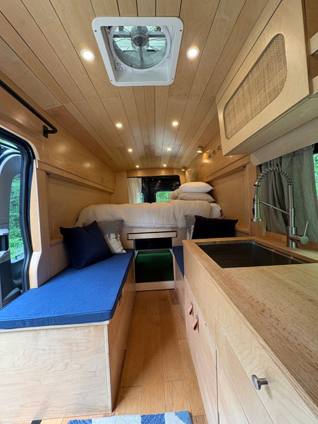 This cozy rv rental interior features warm wood paneling, a comfy bed, kitchen with sink, and blue cushioned seating for travel.