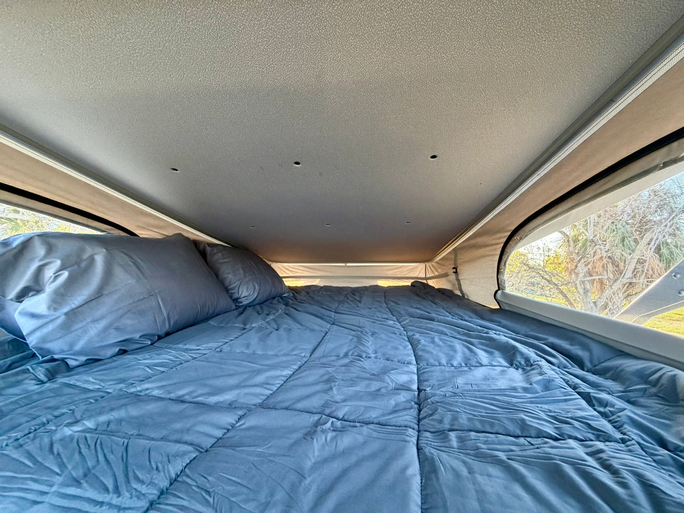 Experience comfort in this RV rental's pop-top bed. A blue comforter and pillows fill the cozy interior, overlooking trees.