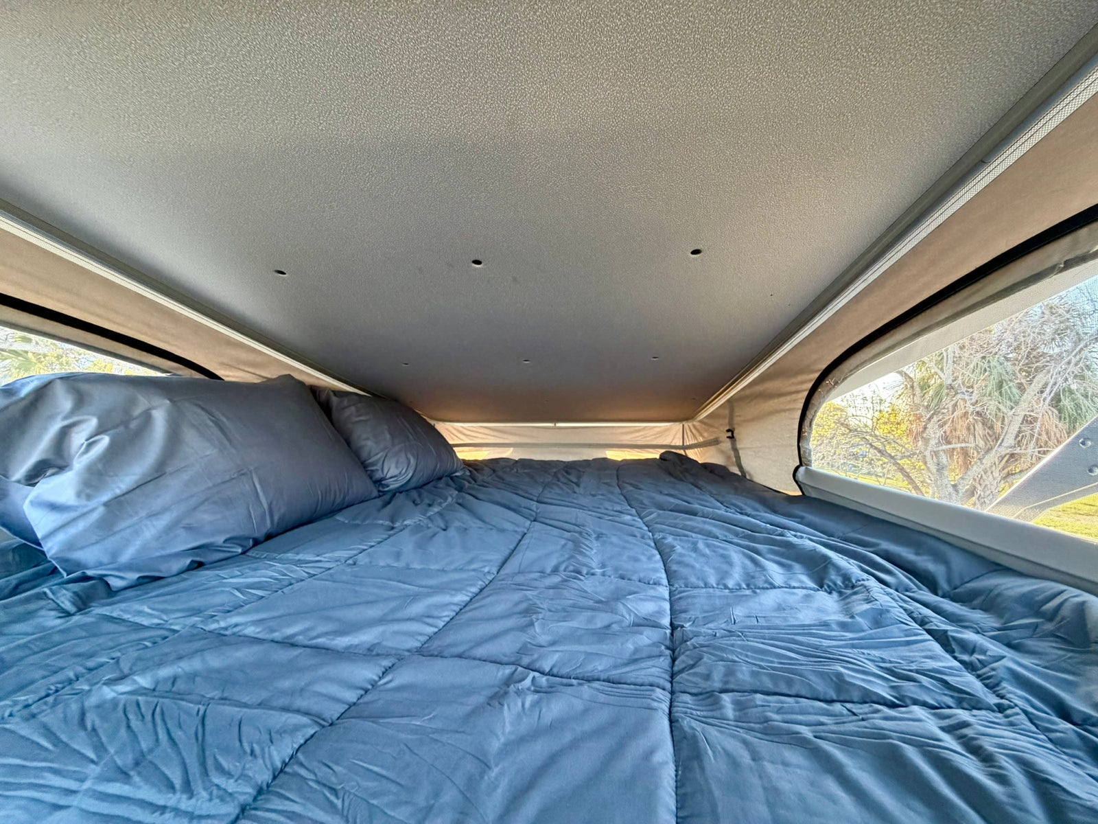 Experience comfort in this RV rental's pop-top bed. A blue comforter and pillows fill the cozy interior, overlooking trees.