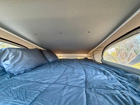 Experience comfort in this RV rental's pop-top bed. A blue comforter and pillows fill the cozy interior, overlooking trees.