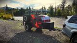 An off-road teardrop trailer, ideal for an RV rental, parked by a river in a scenic mountain setting.
