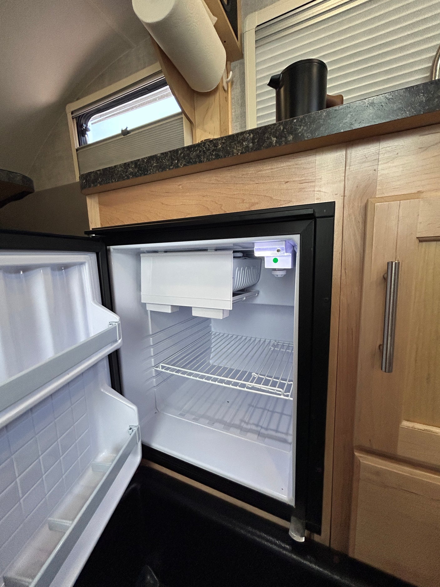 Open, empty mini-fridge in an RV rental, showing compact kitchen with counter, cabinet, and window.