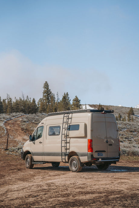 Adventure awaits! Tan Mercedes Sprinter rv rental on a dirt road, amidst trees & snow-dusted hills. Perfect outdoor travel.