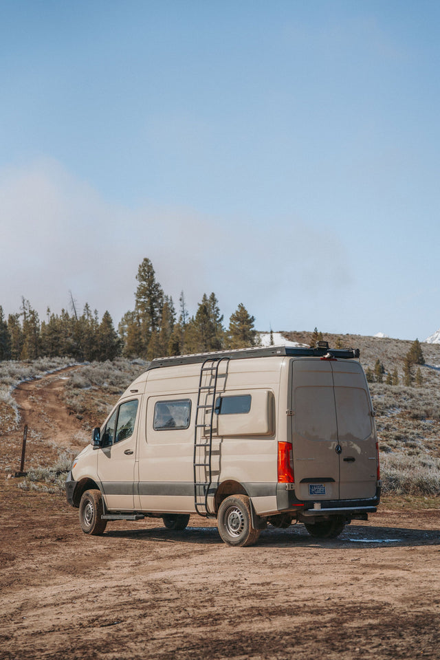 Adventure awaits! Tan Mercedes Sprinter rv rental on a dirt road, amidst trees & snow-dusted hills. Perfect outdoor travel.