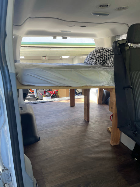 RV rental van's interior: an elevated bed with a white mattress & patterned pillow, offering under-bed storage.