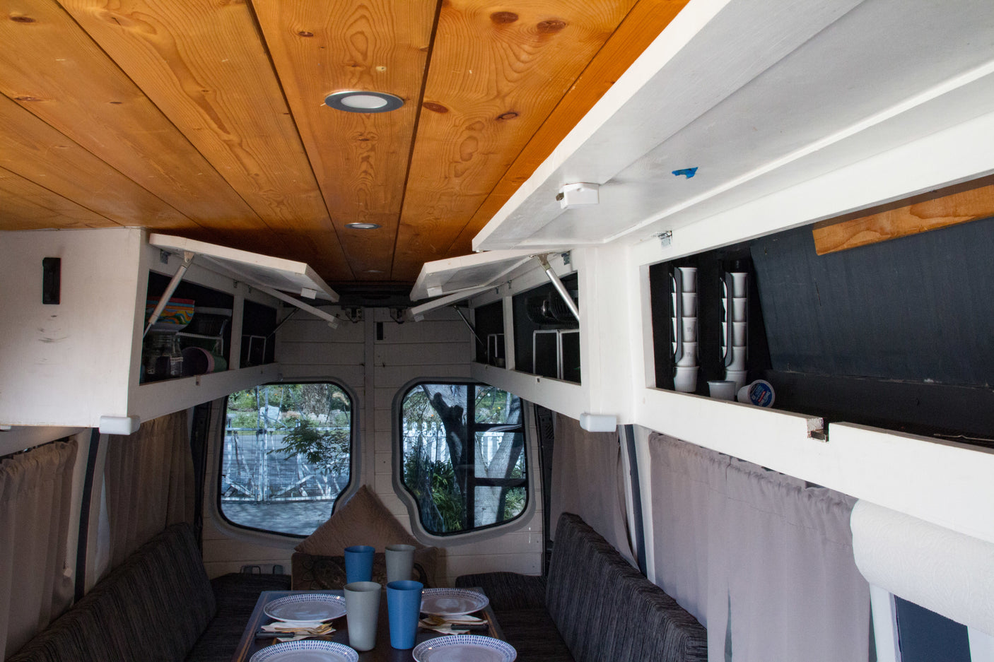Cozy RV rental dining area with a wooden ceiling, open white cabinets, set table with plates & cups, and scenic views.
