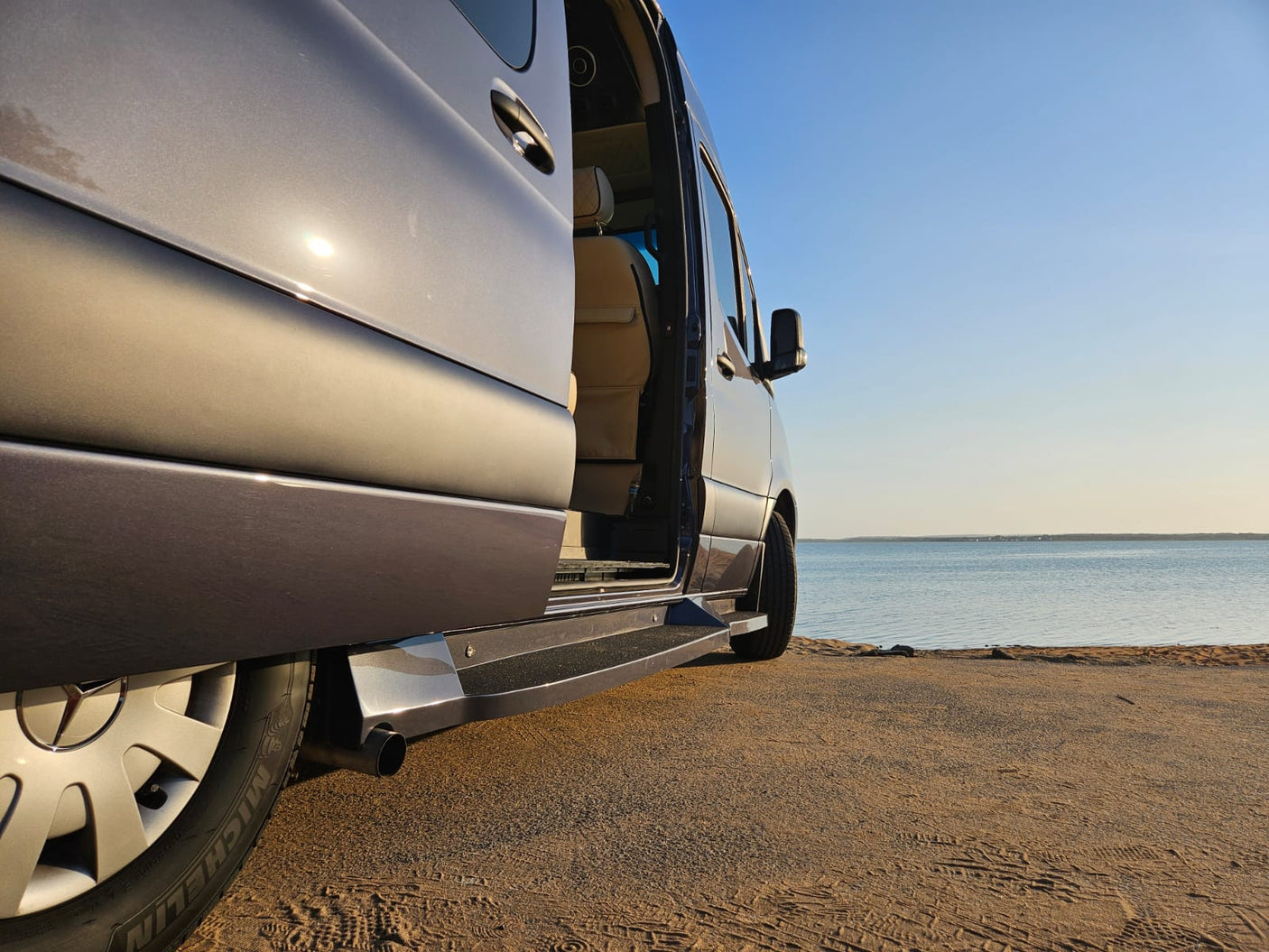 RV rental parked on a sandy beach next to calm water, its side door open for a relaxing view.