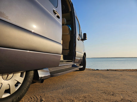RV rental parked on a sandy beach next to calm water, its side door open for a relaxing view.