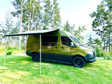 Olive "Vanquest" camper van RV rental with extended awning on green grass. Scenic backdrop of trees and distant mountains.