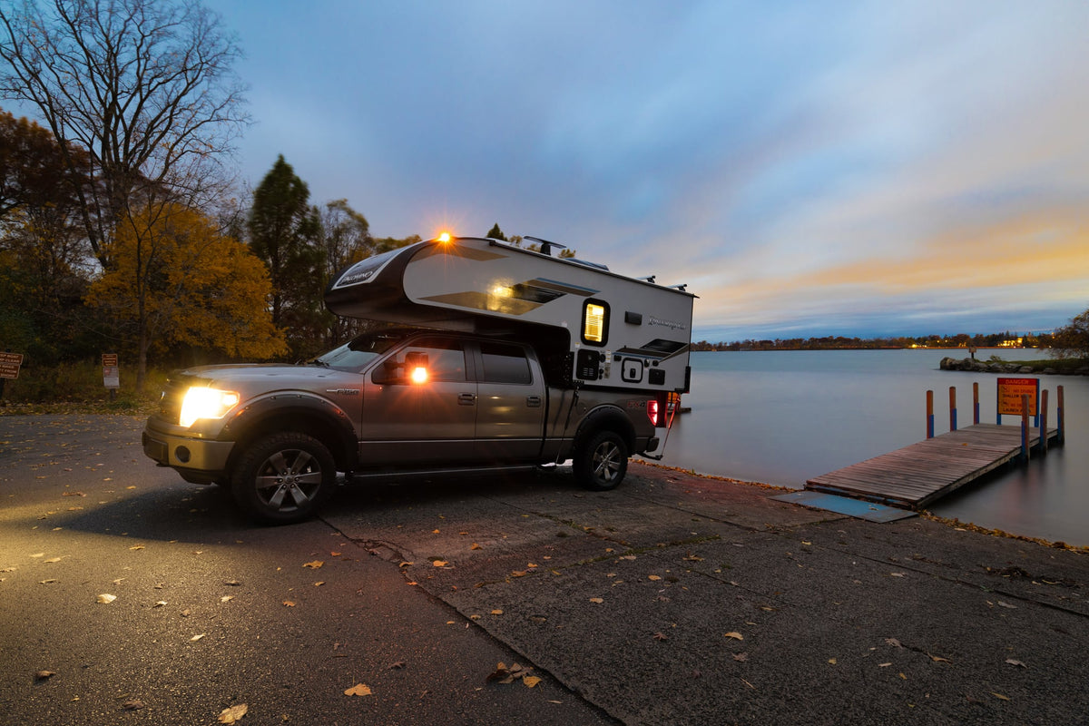 Twilight scene: Ford F-150 with a Palomino truck camper parked at a calm lake's boat launch. Ideal rv rental for outdoor adventures.|Illuminated Palomino truck camper RV rental on a pickup truck