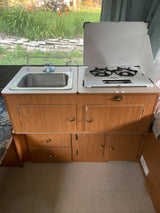 Compact RV rental kitchen features a sink, two-burner stove with cover, and wood cabinets. A scenic outdoor view is visible.